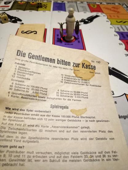 Die erste Seite der Spielregeln mit vereinzelten Flecken (das Spiel ist fast 60 Jahre alt). Dahinter ein Teil des Spielbretts mit dem Meeple, der den Detektiv darstellt. Links und rechts ragen Haken aus ihm raus. Daran baumeln die Geldsäcke, die er uns abgenommen hat.