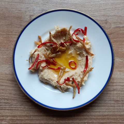 Auf einem kleinen Teller eine kleine Portion Hummus mit Öl in der Mitte + mit frittierten Chili, Ingwer + Knoblauch bestreut