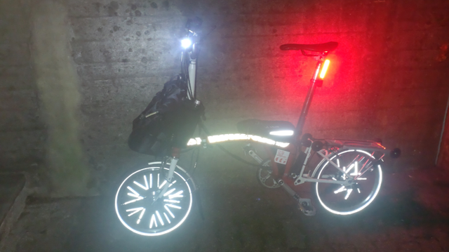 A red and white 16" Brompton H6RD folding bicycle leaning on a concrete wall in the night and photographed with a flash light. Many parts are reflecting the flash light in bright white: The tire walss, the spokes (front wheel more near the rim, rear wheel more near the hub), a line on the frame bag on top of the main tube near the seat post and the upper side of the whole main tube with exception of the folding hinge. Additionally there are bright "be seen" battery lights on on the handlebar and on the seat post directly under the saddle.