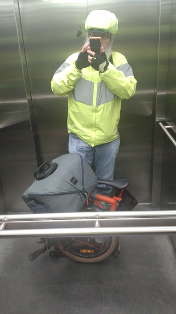 Selfie with folded orange Brompton G-Line with grey front bag in an escalator with mirrors as walls. The Brompton is partially hidden by a metal hand rail. I wear blue jeans, a hi-viz neon-yellow rain jacket with large reflective stripes and a neon-yellow rain cover over the bicycle helmet.