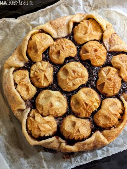 Schwarzes Backblech, helles, zerknittertes Backpapier darauf. Darauf zu sehen eine Apfeltarte, der Belag besteht aus halbierten, braun gebackenen, geschälten Äpfeln, in die Äpfel sind gruselige Gesichter geschnitzt, die durch das Backen zusätzlich gerunzelt sind.