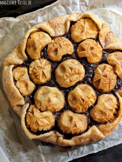 Schwarzes Backblech, helles, zerknittertes Backpapier darauf. Darauf zu sehen eine Apfeltarte, der Belag besteht aus halbierten, braun gebackenen, geschälten Äpfeln, in die Äpfel sind gruselige Gesichter geschnitzt, die durch das Backen zusätzlich gerunzelt sind.