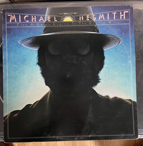 The image features an album cover for "From A Radio Engine to the Photon Wing" by Michael Nesmith. The cover displays a silhouetted figure wearing a hat against a gradient blue background, with the title prominently featured at the top.