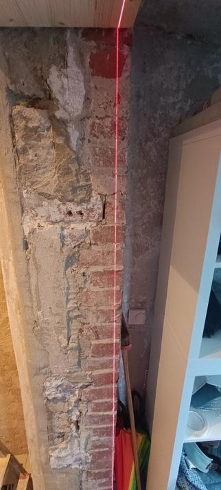 Zentral ist ein gemischtes Mauerwerk das sich in der Bildmitte von unten bis oben erstreckt. Auf dem Mauerwerk leuchtet eine senkrechte Laserlinie die zeigt, dass die Wand ein Neigung nach rechts von 4cm hat.
Rechts schließt sich eine Nische mit einem weißen Ikea Regal an.