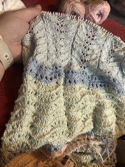 Keskeneräinen huivi pyöröpuikolla. Huivissa on pastellinsävyisiä puuvillalankoja, ja erilaisia osioita yksinkertaisia pinta- ja pitsineuleita. Huivi on mytyssä mutta se on kuunsirpin tai levennetyn puoliympyrän muotoinen.