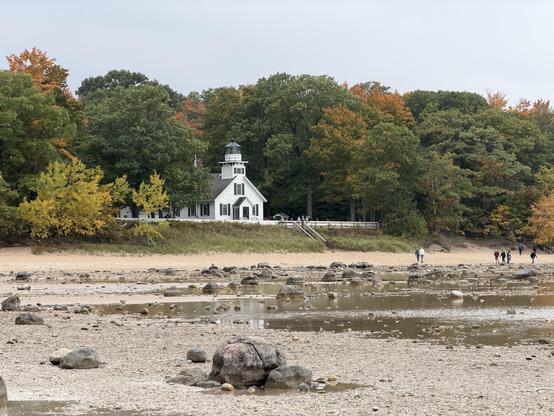 A white lighthouse stands near a sandy beach surrounded by trees displaying autumn colors. The shoreline features scattered rocks, and a few people are visible walking along the beach. The scene is overcast, suggesting a cool day. This is the Old Mission peninsula lighthouse.