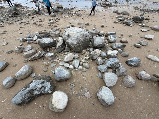 An area of sandy beach scattered with various sizes of rocks and pebbles forming a spiral galaxy sort of design. In the background, a few people are observed, some navigating the rocky terrain.