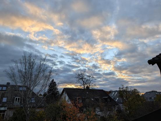 Ciel nuageux laissant passer un peu de ciel bleu. Certains nuages sont éclairés par en dessous par le soleil Ue l'on ne voit pas. Le paysage est composé d'e toits d'immeubles de 3 etages qui émergent entre les arbres aux couleurs d'automne