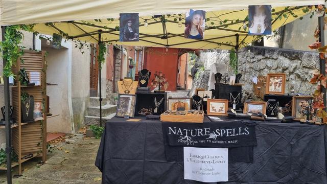 English A stall in a market full of decorations made from dried plants (frames, glass domes) and stained glass jewelry with pressed plants. The stall is decorated with plants and a banner that reads “Nature Spells - Connecting you to nature's magic”.
Français Un stand dans un marché plein de décorations à base de plantes séchées (cadres, cloches en verre) et de bijoux en vitrail avec des plantes pressées. Le stand est décoré de végétaux et une bannière dit "Nature Spells - Connecting you to nature's magic".