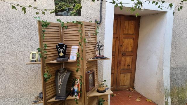 English A stall in a market with a shelf full of decorations made from dried plants (frames, glass domes) and stained glass jewelry with pressed plants. The stall is decorated with plants.
Français Un stand dans un marché avec une étagère pleine de décorations à base de plantes séchées (cadres, cloches en verre) et de bijoux en vitrail avec des plantes pressées. Le stand est décoré de végétaux.