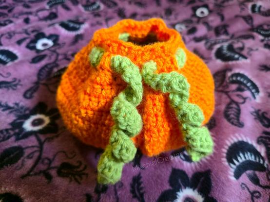 A crochet dice bag that looks like a pumpkin, with curly green drawstrings that look like vines.