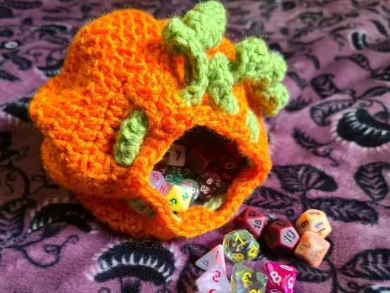 A crochet dice bag that looks like a pumpkin, with curly green drawstrings that look like vines. There are dice spilling out of the bag.