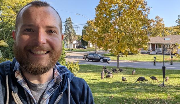 A selfie of a bearded man (me), with a bunch (10/11?) of turkeys in the background near a bird feeder of a suburban yard.
