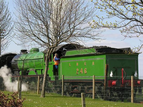 LNER 246 "Morayshire" approaches Bo’ness station. The image shows a light green steam train with a visible part of a carriage attached to its front, left-hand end and a driver leaning out as the engine reverses to the right. There are trees that are not in leaf this side of the railway line with a fence and grass. The sky is light blue.