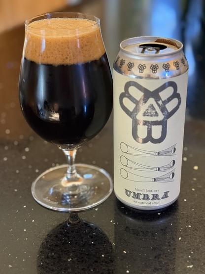 A snifter glass full of dark beer with a bubbly dark tan head. The pint can has a light tan label with silver insignia and lettering depicting the shadows of a lunar eclipse on the earth