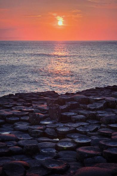 A vertical photograph of the Giant's Causeway at sunset. The foreground is dominated by the dark, hexagonal basalt columns, some with pools of water reflecting the light. The sea stretches out to the horizon in the middle ground under a dramatic, vibrant orange and pink sky where the bright sun is low, casting a shimmering reflection across the water towards the shore. The atmosphere is tranquil and majestic.
