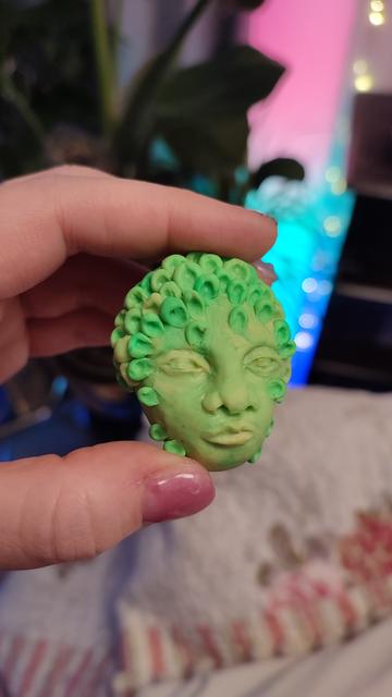 Holding a polymer clay head, ca. 8cm high. It's light green with some swirls of lighter and darker shades. Neutral expression, I'd say. Forehead is covered in a repeating pattern of little... Ears? Or rolled up leaves? They spread to the side, some even down to the chin. It's like leaves overgrowing the head or mushrooms. They're of the same clay, so they must be part of the organism.