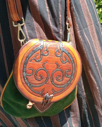 Close-up of a sporran-shaped purse, the round lid tooled and painted as a hobbit door, different shades of brown for the wood planks, dark gray metallic painting for the metal decorations, nails and button, and darkened brown border around, green suede for the body of the bag and dark yellow bias. Beige thread and golden snap hooks attached to a brown shoulder strap