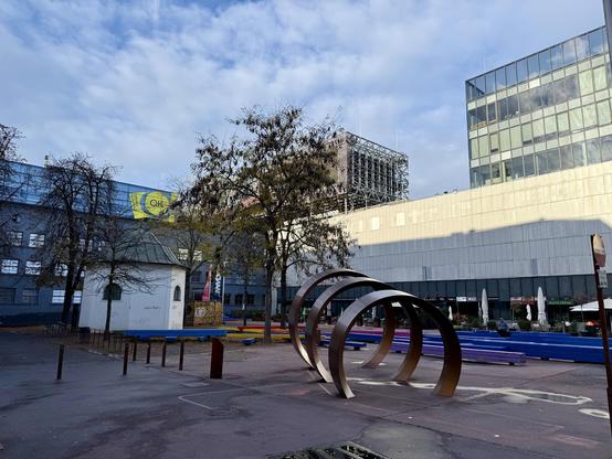 Ein Platz, auf dem ein Kunstwerk und einige Bäune stehen, und der von modernen Gebäuden umgeben ist. Vor dem rechten Gebäude sind Anzeichen von Außengastronomie zu erkennen.