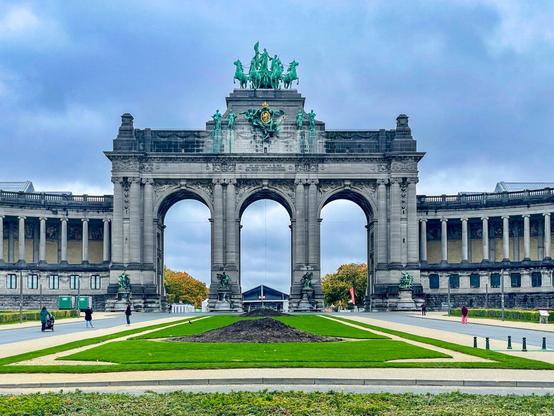 Photo of the Cinquantenaire Arch in Brussels, Belgium — a grand stone monument with three arches topped by a bronze sculpture of horses and figures, framed by green lawns and a cloudy blue sky with people walking nearby.