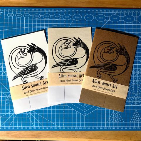 a set of three cards laid out on a blue cutting mat. a brown Kraft card, a plain white card, and a cream parchment pattered card. each card has a yellow parchment strip of paper wrapped around it that says "Alien Sunset Art" Hand Block Printed Card.

the block print on the cards is of  a creature with two heads, wings and two legs with paws, one head is situated at the end of a long tail that is forming a spiraling circle up and over the main body, the ’main’ Head is biting the tail as it circles near it and the second head is looking at it in shock/outrage/despair