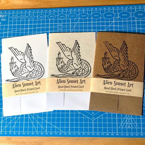 a set of three cards laid out on a blue cutting mat. a brown Kraft card, a plain white card, and a cream parchment pattered card. each card has a yellow parchment strip of paper wrapped around it that says "Alien Sunset Art" Hand Block Printed Card.

the block print on the cards is of  a a dragon standing it’s own tail like a platform spouting fire straight up with unfurled feathery wings