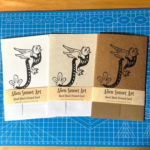 a set of three cards laid out on a blue cutting mat. a brown Kraft card, a plain white card, and a cream parchment pattered card. each card has a yellow parchment strip of paper wrapped around it that says "Alien Sunset Art" Hand Block Printed Card.

the block print on the cards is of  a flying snake like creature with feathery wings, a large leaf like structure at the end of their tail and a round furry head and sharp teeth.