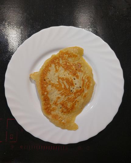 Ein goldbraun gebratener Pfannkuchen liegt auf einem weißen, gewellten Teller. Der Pfannkuchen ist leicht unregelmäßig geformt, hat aber eine appetitliche Farbe und sieht fluffig aus. Der Teller steht auf einer schwarzen Herdplatte.