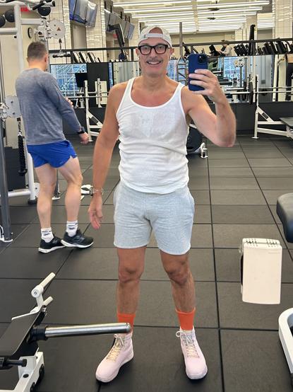 A man (me) wearing a white tank top, gray shorts, and pink sneakers with orange socks stands in a gym, taking a selfie. In the background, another person is partially visible, walking by. The gym is well-equipped, with various machines and mirrors