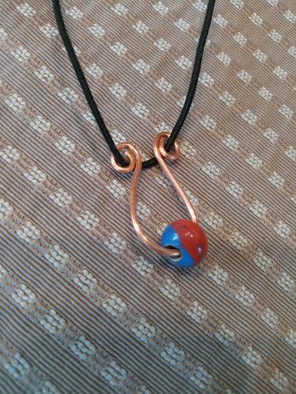 Auf einem Deckchen liegt ein aus Kupferdraht gebogener Anhänger mit einer blauroten Glasperle, aufgefädelt auf schwarzes Lederband.
