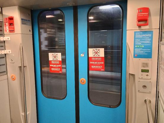 Foto von Zugtür mit "defekt"-Aufklebern, im Hintergrund Bahnsteig.