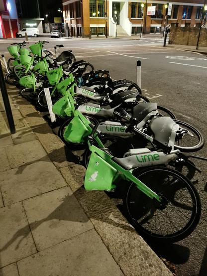 A number of green and white Lime bikes at the side of a road, most of them are on the ground. Looks like one had been pushed over which caused the others to fall.