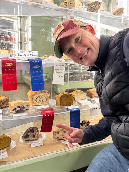Picture of me (middle aged white guy in a puffy black vest and a hat with the state flag of North Carolina on it, pointing excitedly to the second place ribbon. The exhibits are all in a case and you see lots of breads in the case. My half loaf of bread is on the plate and you can see the spiral of the cinnamon filling that I rolled up into a loaf.