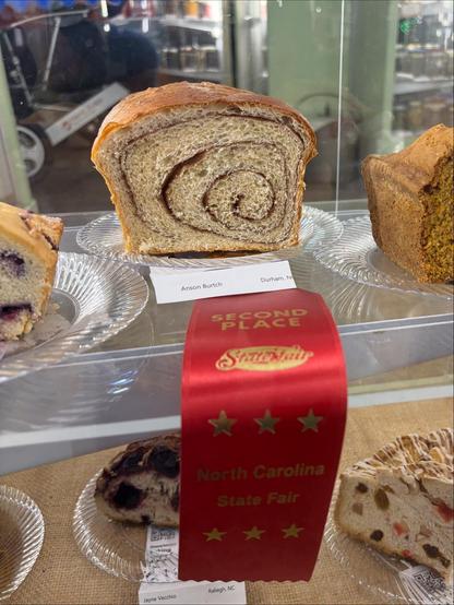 Half loaf of white bread cut in half. You can see the swirl of the cinnamon filling that has been rolled up into a loaf and baked. There is a red second place ribbon hanging from the plate.