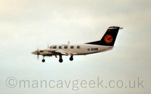 Slightly fuzzy side view of a twin engined BizProp flying from right to left at a low altitude, with undercarriage lowered and flaps extended from the rear of the wings, suggesting it is about to land.
The plane is largely white, with small black "Lufthansa" titles on the upper fuselage, large black registration "D-IOSB" on the rear fuselage, and a dark blue tail with a yellow circle containing a stylised blue flying bird.
Flat grey sky fills the rest of the frame.