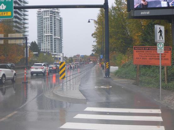 Multi-use path along Marine Drive (looking east). The road and path are wet from rain. Photo taken through the construction fence.