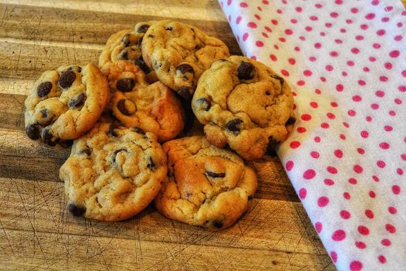 The image shows a pile of chocolate chip cookies on a wooden surface. The cookies are stacked somewhat haphazardly and overlap each other. Each cookie has a slightly irregular shape and is covered in chocolate chips.