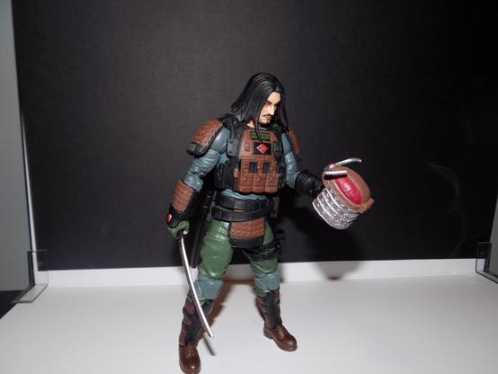 A man in a hybrid of modern armor and samurai armor looks down at his helmet, sword in the other hand