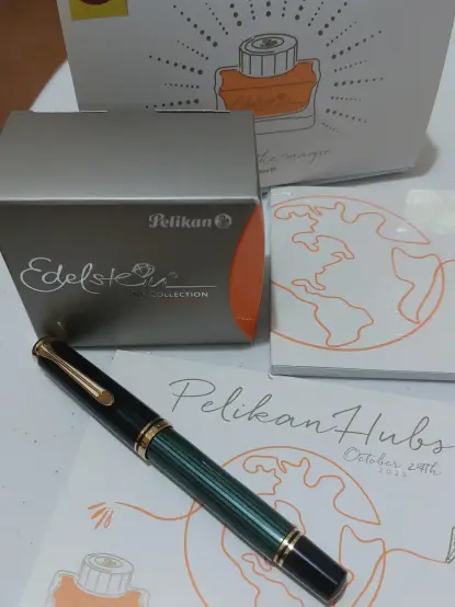 A Pelikan M600 green-striped fountain pen, next to a box of Pelikan Edelstein Mandarin ink, a notepad and a postcard that says Pelikan Hubs, October 24, 2025.