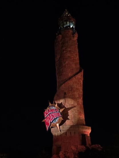 A photograph of the Pharos Lighthouse located outside of Universal Islands of Adventure in Orlando, Florida. It looks to be made of stone with a spiraling staircase going up the exterior. There is a large sign that says "UNIVERSAL'S ISLANDS OF ADVENTURE", with "UNIVERSAL'S" being much smaller compared to the name of the park. This sign is illuminated by spotlight. The lighthouse stands out against the black night sky.