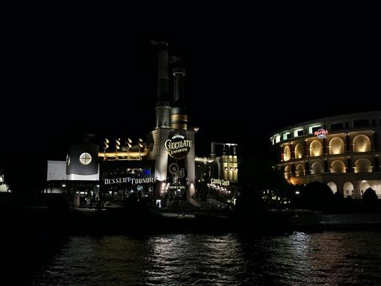 A photograph of The Toothsome Chocolate Emporium at Universal CityWalk Orlando, taken at night across from a body of water. The Chocolate Emporium resembles a steampunk-styled factory and is located next to a coliseum-looking building, which is the Hard Rock Cafe. The lights from the buildings is being reflected by water, making the ripples visible.