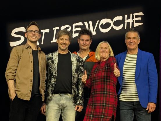 Bild aller Teilnehmer auf der Bühne. Alle lächeln in das Publikum. PE hat beide Daumen gehoben. 
Im Hintergrund steht Satirewochen.