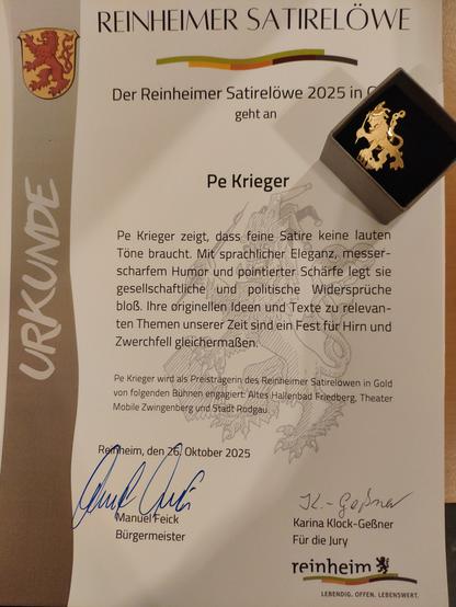 Foto der Urkunde und des kleinen goldenen Löwens. 
Auf der Urkunde steht: 
Pe Krieger zeigt, dass feine Satire keine lauten Töne braucht. 
Mit sprachlicher Eleganz, messerscharfen Humor und pointierter Schärfe legt sie gesellschaftliche Widersprüche bloß.
Ihre originellen Ideen und Texte zu relevanten Themen unserer Zeit  sind ein Fest für Hirn und Zwerchfell gleichermaßen.