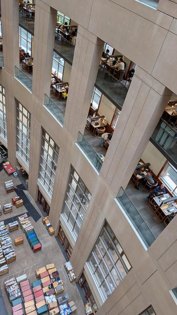 Inside the Vancouver Public Library. Picture taken from an upper floor looking down at the curved side wall galleries. People sitting at desks, working. All the way down is a non-public space filled with dozens of carts filled with books to be sorted. A great sense of height comes out of the picture.