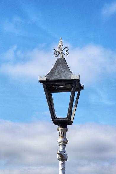 A vintage-style, black metal street lamp set against a bright blue sky with scattered white clouds. The lamp features a square, glass-enclosed lantern with a peaked roof, topped by an ornate metal finial. The pole is cylindrical, painted in a light grey colour, and shows signs of weathering, adding to its antique charm. There is no lightbulb inside the lamp.