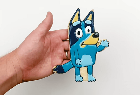A hand holds a decorated sugar cookie shaped like Bluey, the blue heeler dog character, with royal icing details showing her distinctive blue coat, yellow inner ears, and cheerful expression.