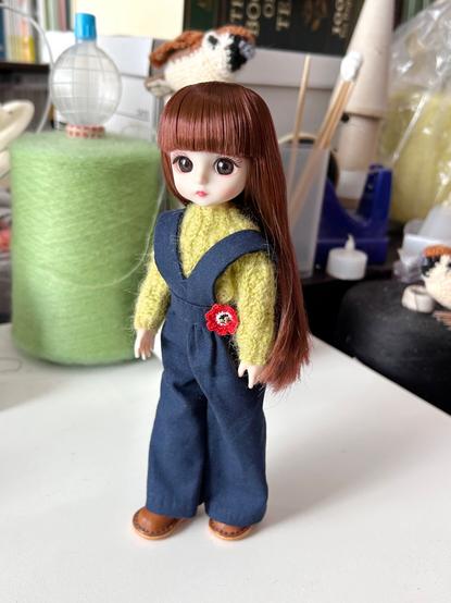 長い茶色の髪をした人形が、黄緑色のセーターと紺色のオーバーオールを着て立っています。オーバーオールには赤とオレンジ色のクロシェ編みの花が飾られており、茶色い革のブーツを履いています。背景には緑色の糸巻きなど、手芸用品が見えます。
English:
A doll with long brown hair stands wearing a fuzzy green-yellow sweater and navy blue overalls, which are decorated with a red and orange crocheted flower. The doll is wearing brown leather boots, and craft supplies like a spool of green yarn are in the background.