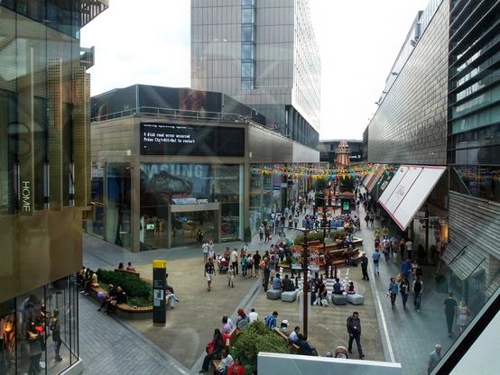A bustling urban shopping street framed by modern glass-fronted buildings, featuring a large digital billboard displaying an error message: ‘A disk read error occurred. Press Ctrl+Alt+Del to restart.’ Below, the street is lively with people walking, sitting, and socialising. Colourful triangular bunting spans overhead, adding vibrancy to the scene. Various stores and brands line the street, contributing to the dynamic and contemporary atmosphere.