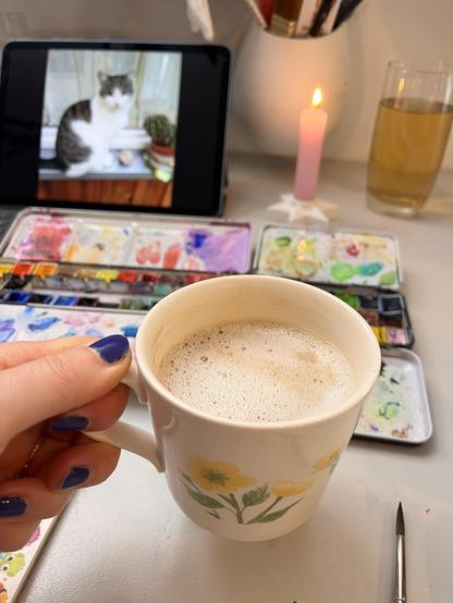 Ich halte eine Tasse mit Cappuccino in der linken Hand. Im Hintergrund sieht man aufgeklappte Malkästen auf dem Tisch, eine brennende Kerze und ein Tablet auf dem unscharf ein Foto einer grau-weißen Katze zu sehen ist.