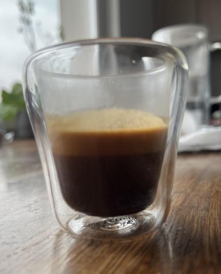 Zbliżenie szklanego kubka do espresso zawierającego warstwy espresso i kremu. Kubek znajduje się na drewnianej powierzchni, z rozmytym tłem pokazującym więcej szkła i zieleni.